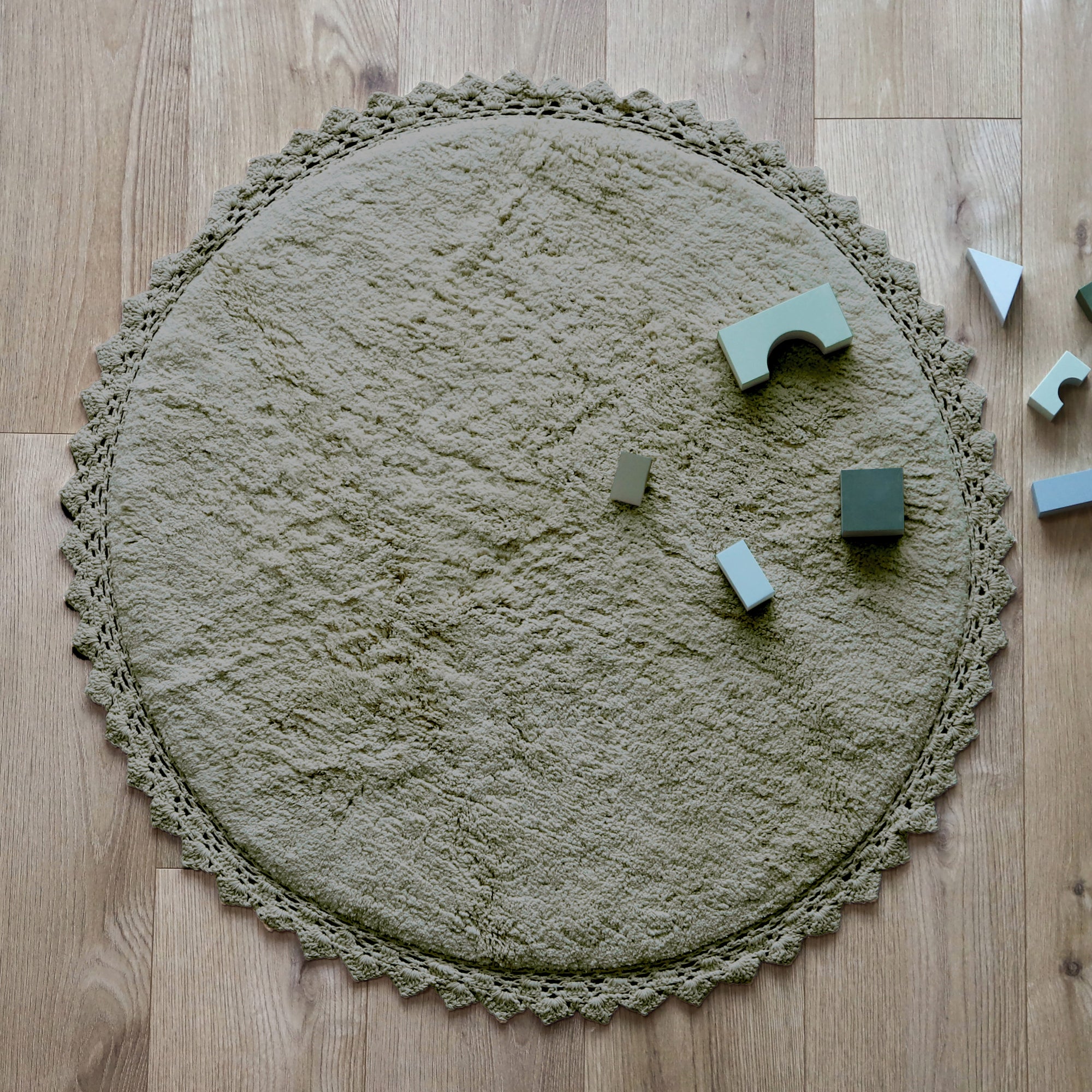 Rond olijfgroen vloerkleed met zachte, organische textuur en gehaakte rand, gelegen op houten vloer met houten blokken erop. - Perla Olive Vloerkleed Nattiot - Default Title - boho, nattiot, perla, perla olive, rond, Vloerkleed - Casa Mini